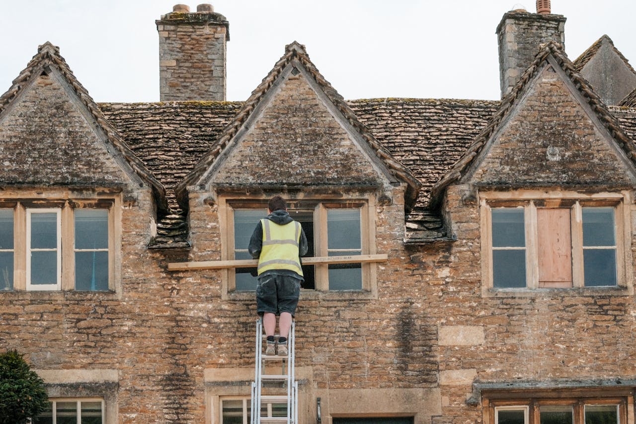 Home A construction worker on a ladder repairs a window on a stone building.